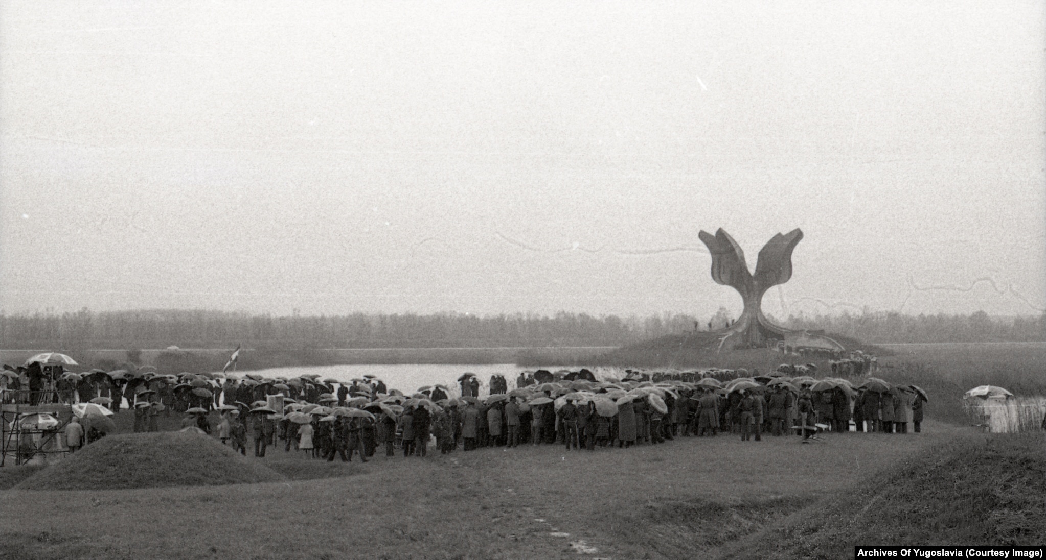 A large crowd with umbrellas standing in rain before the Jasenovac memorial flower, 1980.