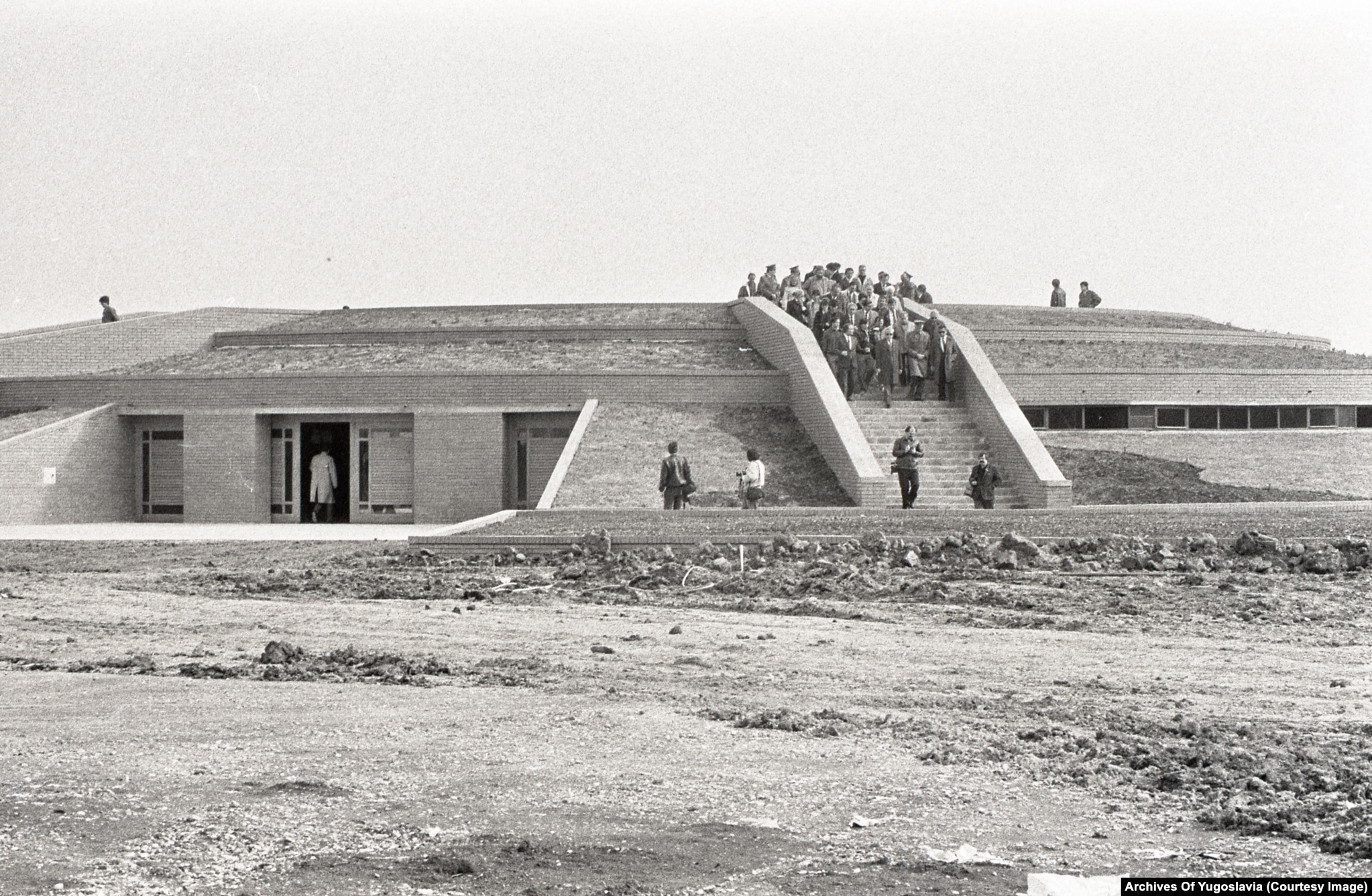 Officials in suits walking along the curved exterior wall of the Sremski Front memorial, 1988.