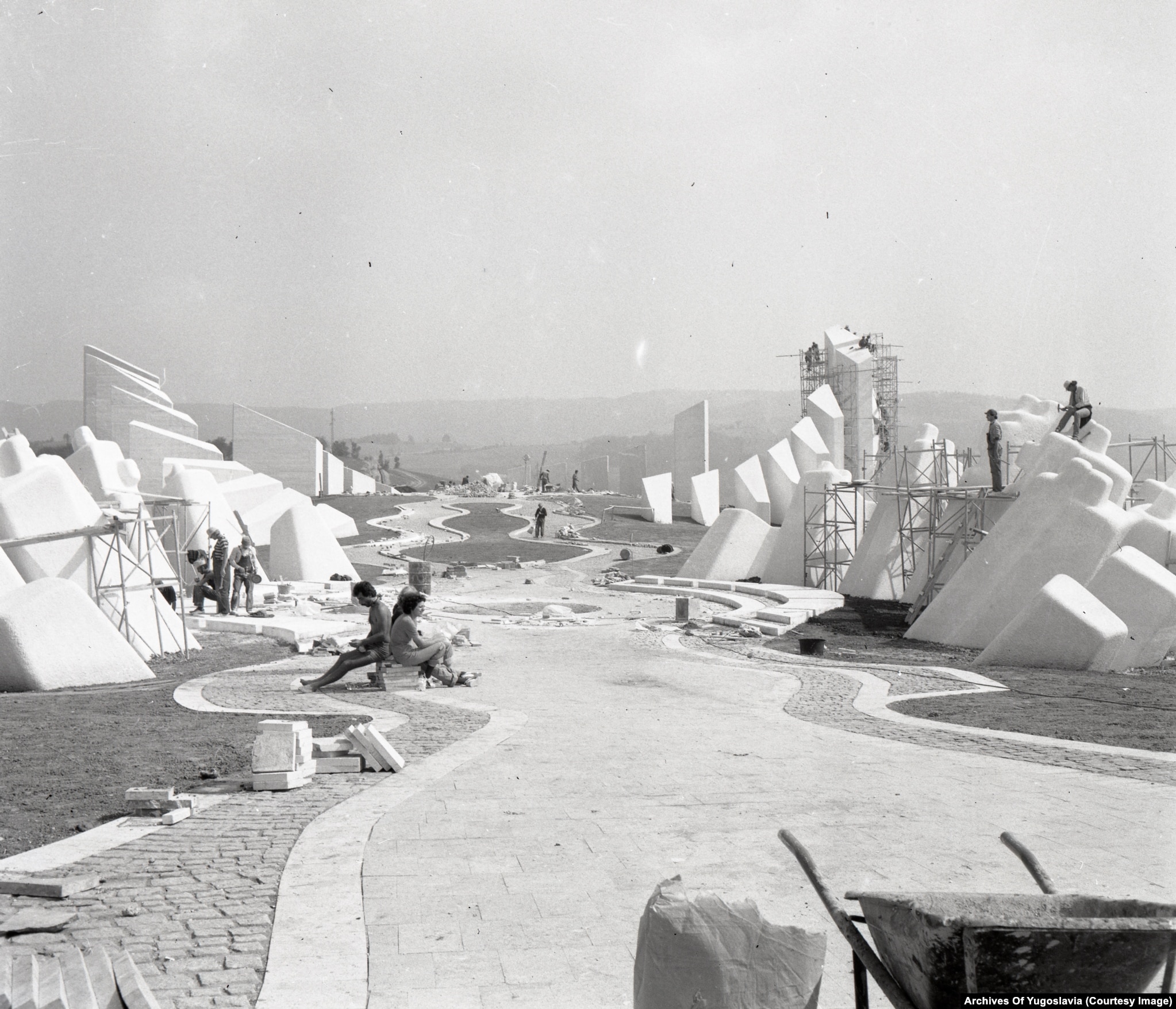 Black-and-white photograph of cranes and unfinished concrete forms on a wooded hilltop, Kadinjača, 1979.